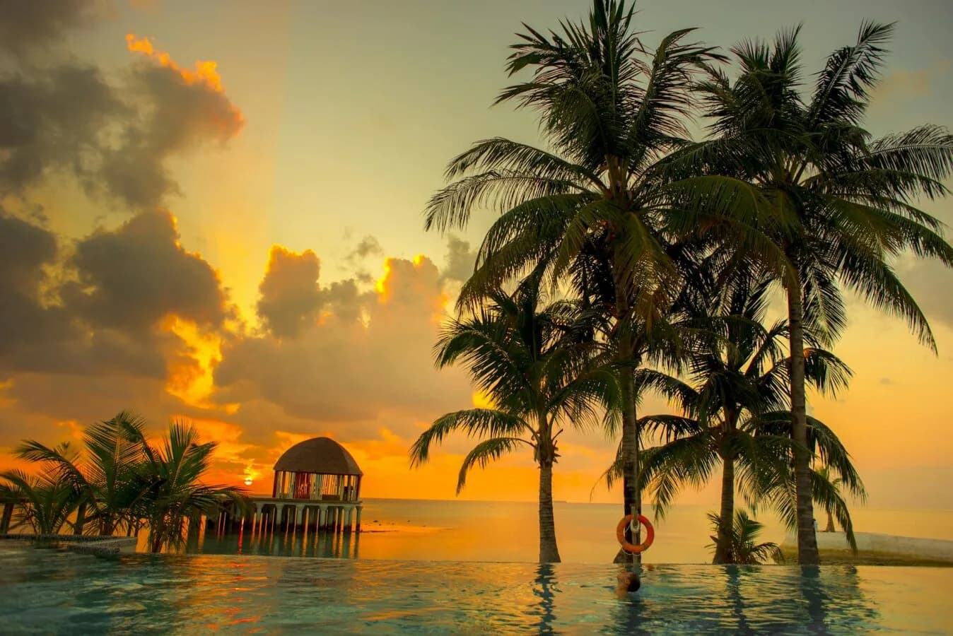 Malediven Sonnenuntergang über dem Meer mit Palmen, Überwasser-Pavillon und infinity Pool im Vordergrund