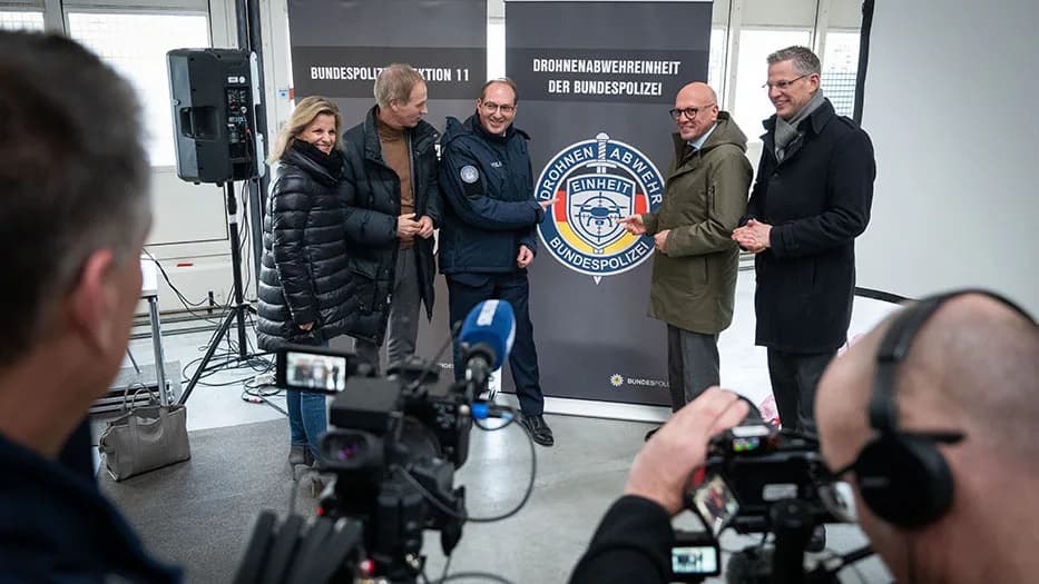 Beamte und Politiker bei Präsentation der Drohnenabwehr-Einheit der Bundespolizei vor Banner mit Emblem, Kameras im Vordergrund