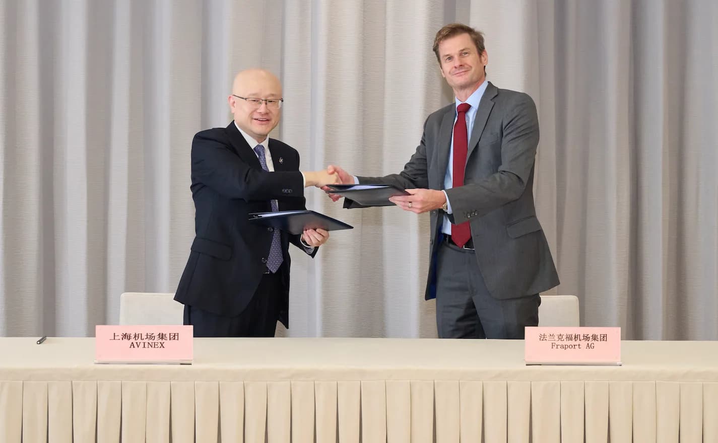 Zwei Geschäftsleute schütteln sich die Hand beim Unterzeichnen von Verträgen an einem Tisch mit Namensschildern Pudong Avinex und Fraport