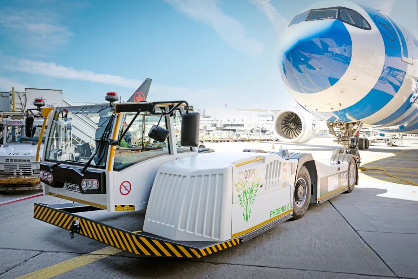 Flugzeugschlepper EcoTow11 am Flughafen neben Passagierflugzeug mit sichtbarem Triebwerk und Bugfahrwerk, Bodenpersonal und Vorfeldfahrzeuge im Hintergrund