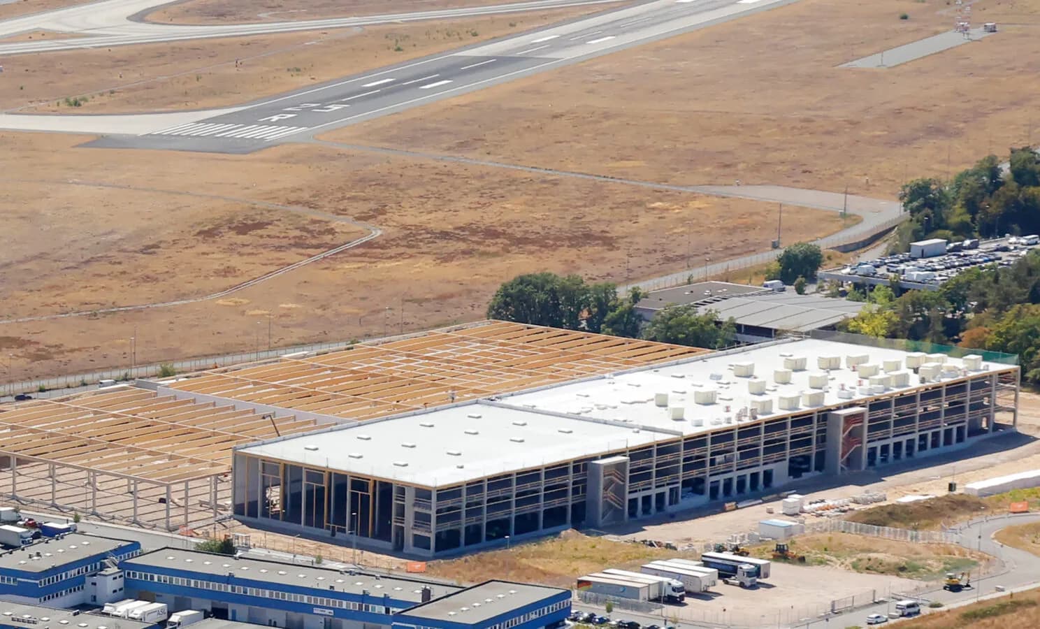 Luftaufnahme LogiSpace Taubengrund Logistikhalle im Bau mit Ladetoren, Lkw und Parkplätzen neben Rollbahn eines Flughafens