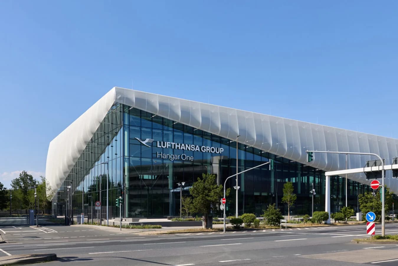 Lufthansa Group Hangar One moderner Glasbau mit geschwungener Fassade, Firmenlogo und ruhiger Straßenfront bei blauem Himmel
