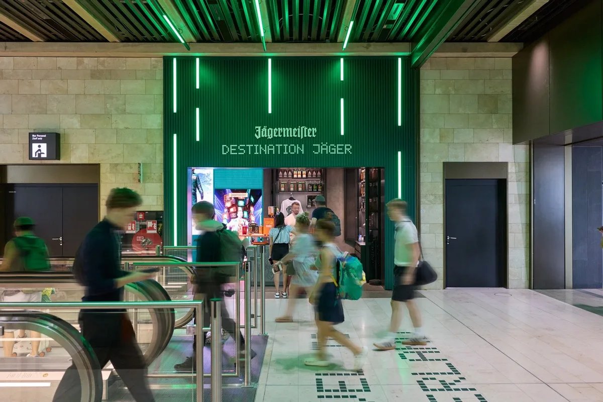 Jägermeister Verkaufsstand "Destination Jäger" mit grüner Neonbeleuchtung in Bahnhofshalle, Passanten und Reisende am Kiosk beim Einkaufen