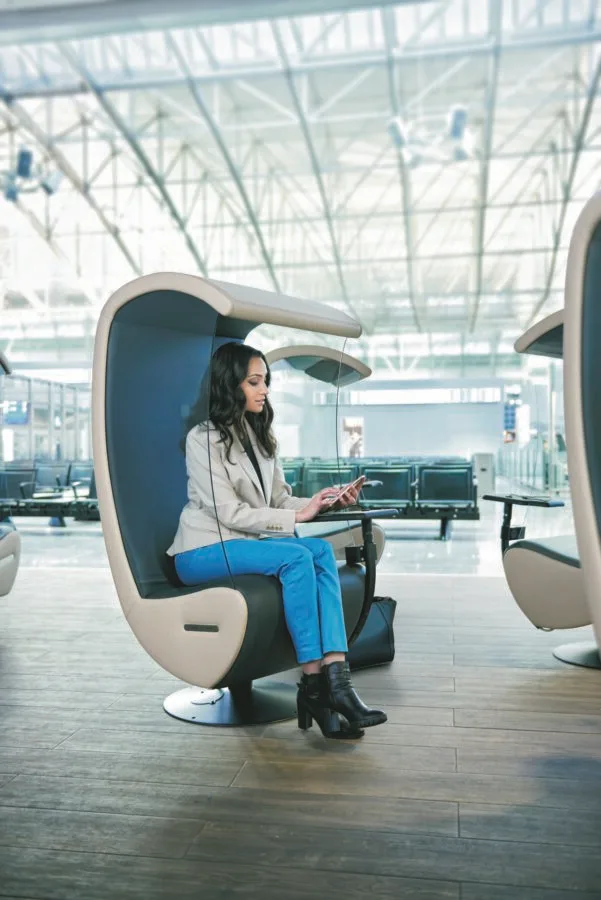 Frau sitzt in moderner Silent Chair Kabine im Flughafen und arbeitet am Tablet, Reisetasche am Boden