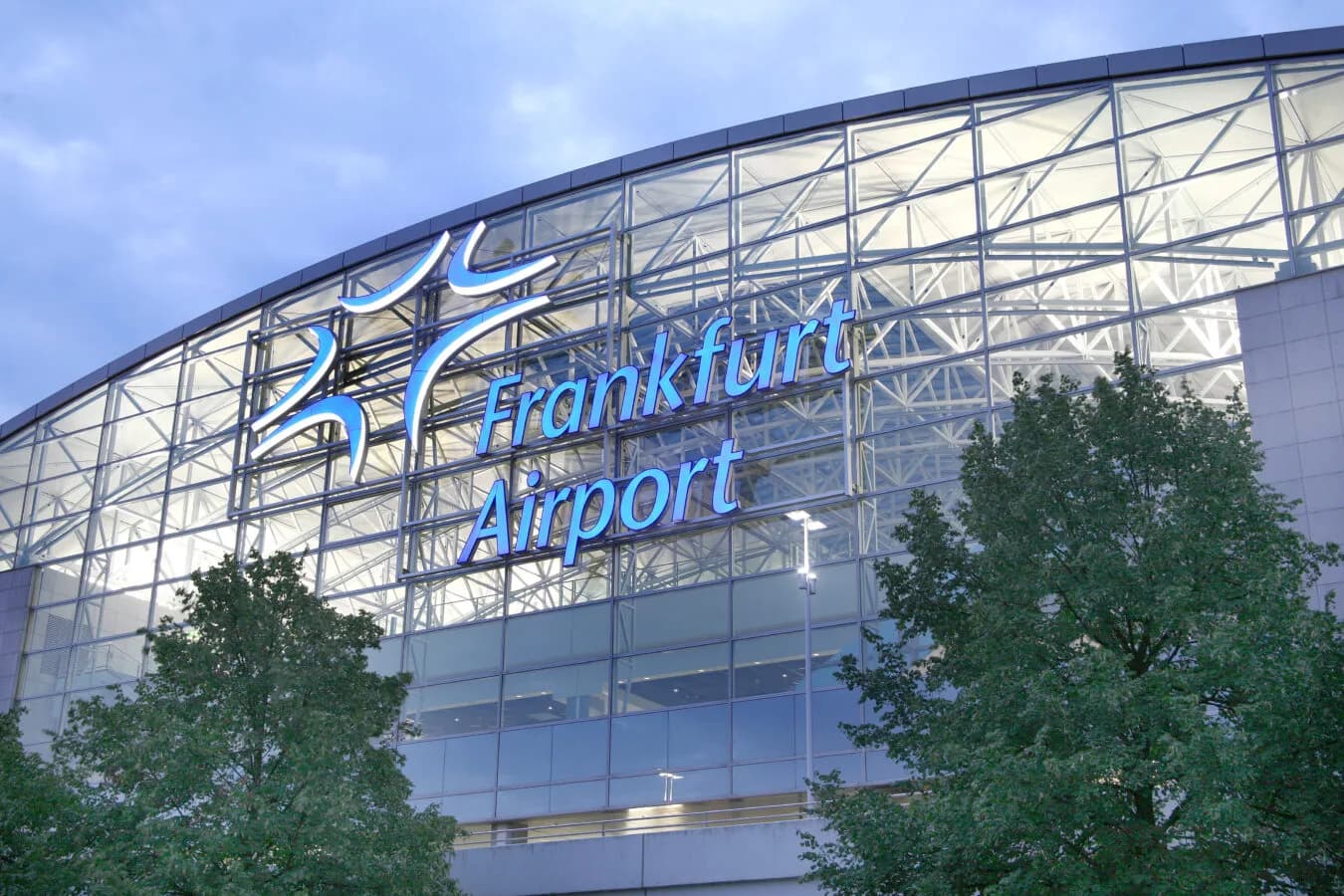 Außenansicht Terminal mit großem beleuchteten Frankfurt Airport Schriftzug und Glasfassade bei Abendlicht, Bäume im Vordergrund