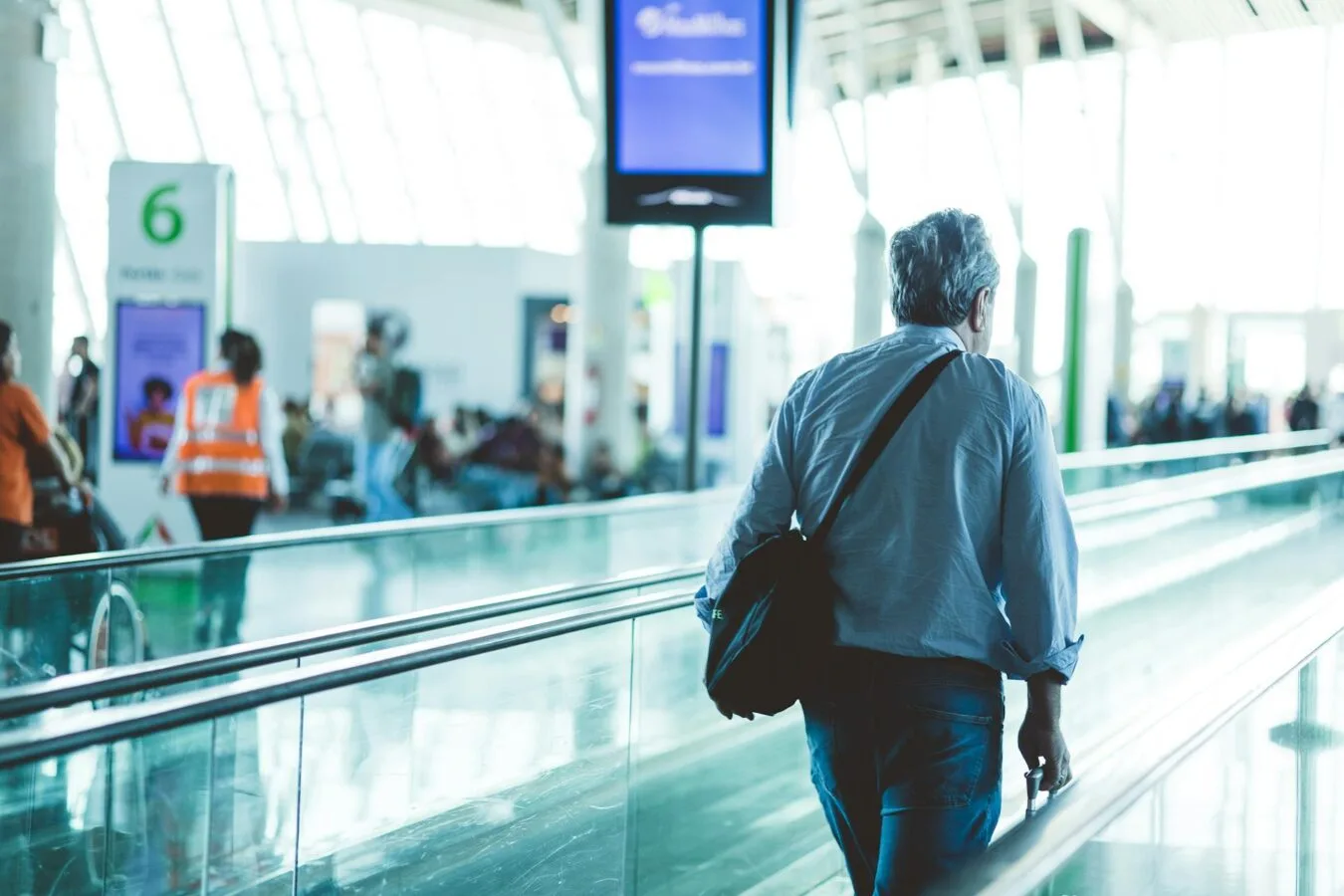 Ălterer Mann mit Schultertasche auf Laufband im Flughafen, Reisender in Abflughalle nahe GepĂ€ckaufbewahrung und Informationsanzeigen