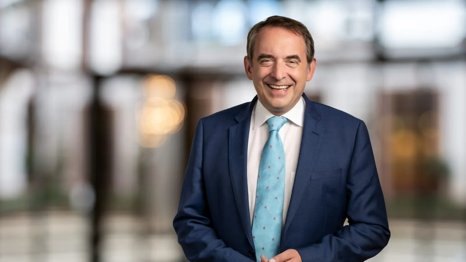 Mann im Anzug lächelt in Büro- oder Lobby-Umgebung, blaues Sakko, hellblau gemusterte Krawatte, formelles Porträt von Prof. Dr. Alexander Lorz