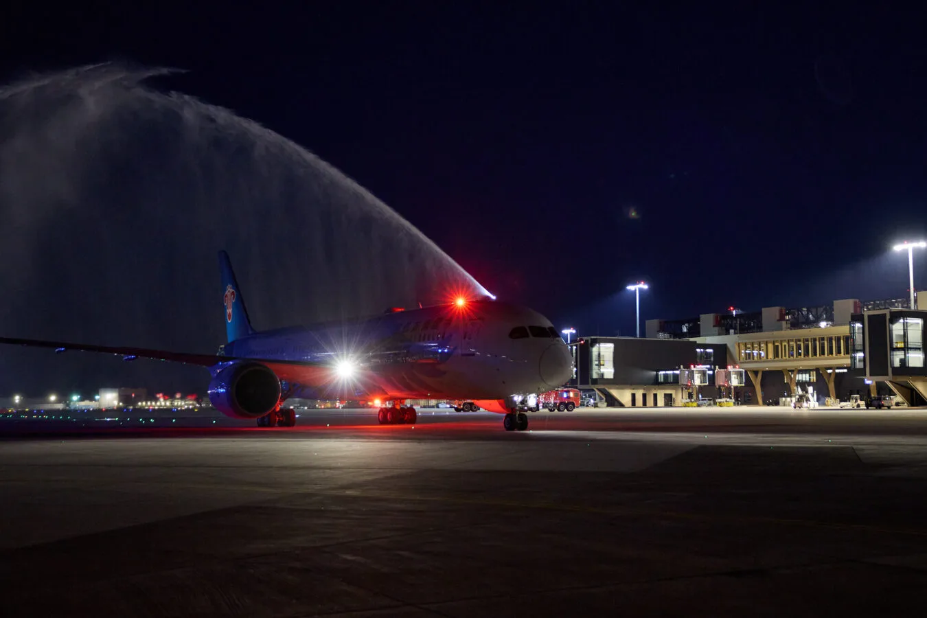 Nachtaufnahme eines Passagierflugzeugs bei der Taufe mit Wasserfontäne am Flughafen, beleuchtete Fluggastbrücken im Hintergrund