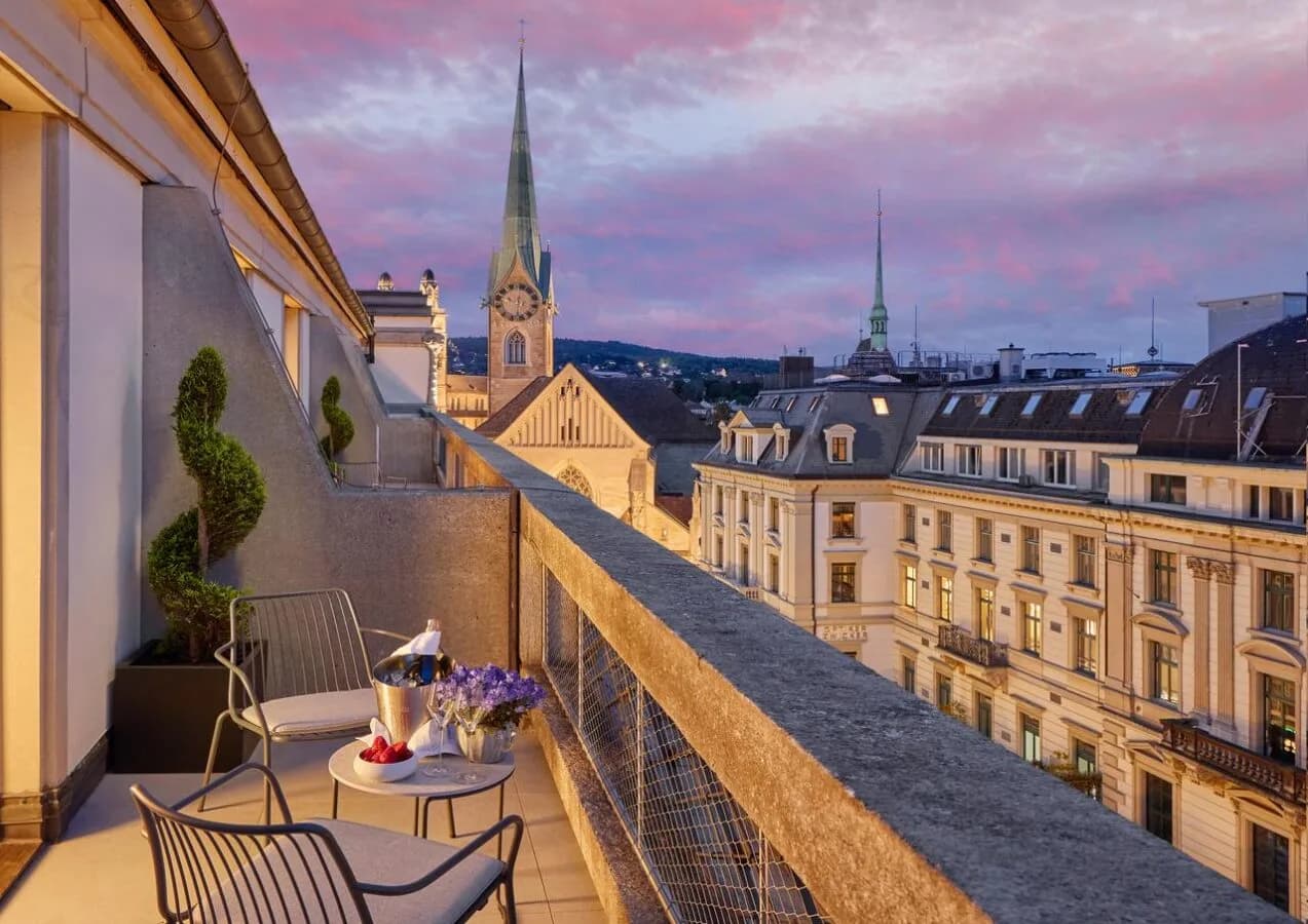 Deluxe Terrassenzimmer Mandarin Oriental Zürich Savoy mit Balkonbestuhlung, Champagner auf Tisch, Blick auf historische Stadtfassaden und Kirchturm bei Abenddämmerung
