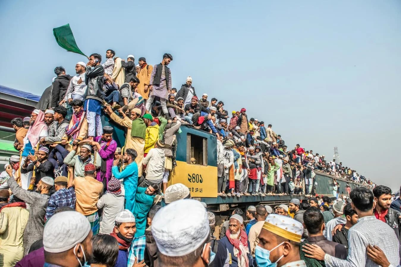 Überfüllter Zug mit Dutzenden Menschen auf Dach und Wagen, dichtgedrängte Pilger in traditioneller Kleidung und Kopfbedeckungen bei chaotischer Abreise am Bahnhof (Flughafenchaos Frankfurt erwähnt)