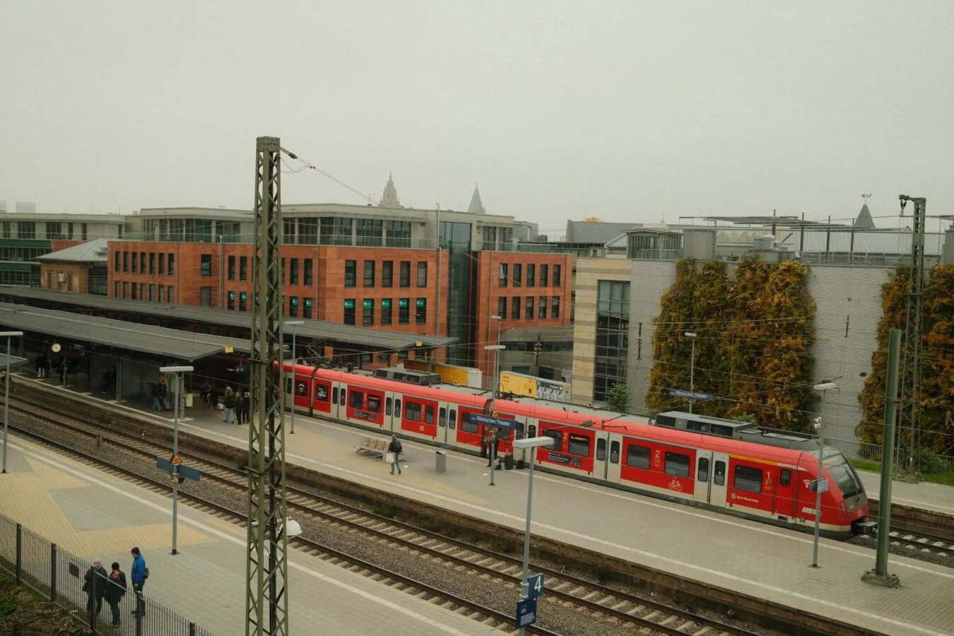 S-Bahn am Bahnsteig des Bahnhofs Mainz mit wartenden Fahrgästen und roten Regionalzügen vor Bürogebäuden
