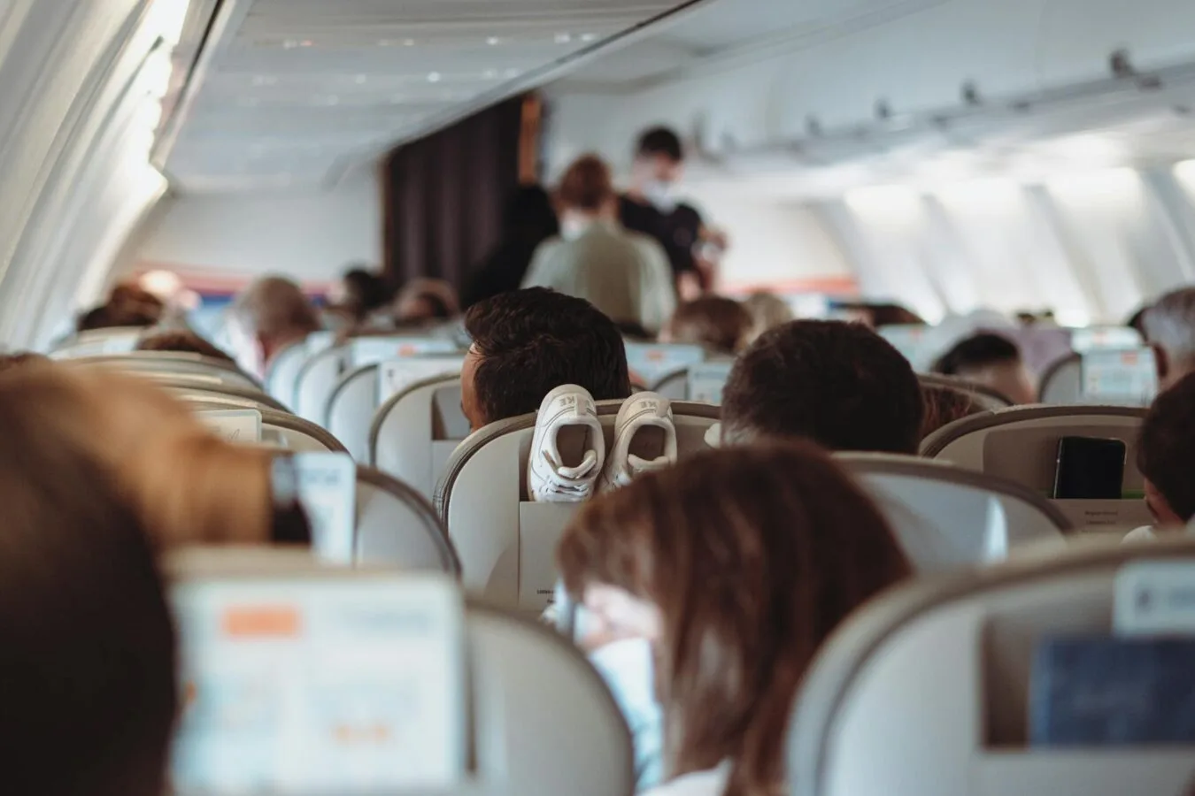 Kabinenansicht eines Flugzeugs mit Reihen von Passagiersitzen, Fluggästen und einer Flugbegleiterin, die Passagiere überprüft