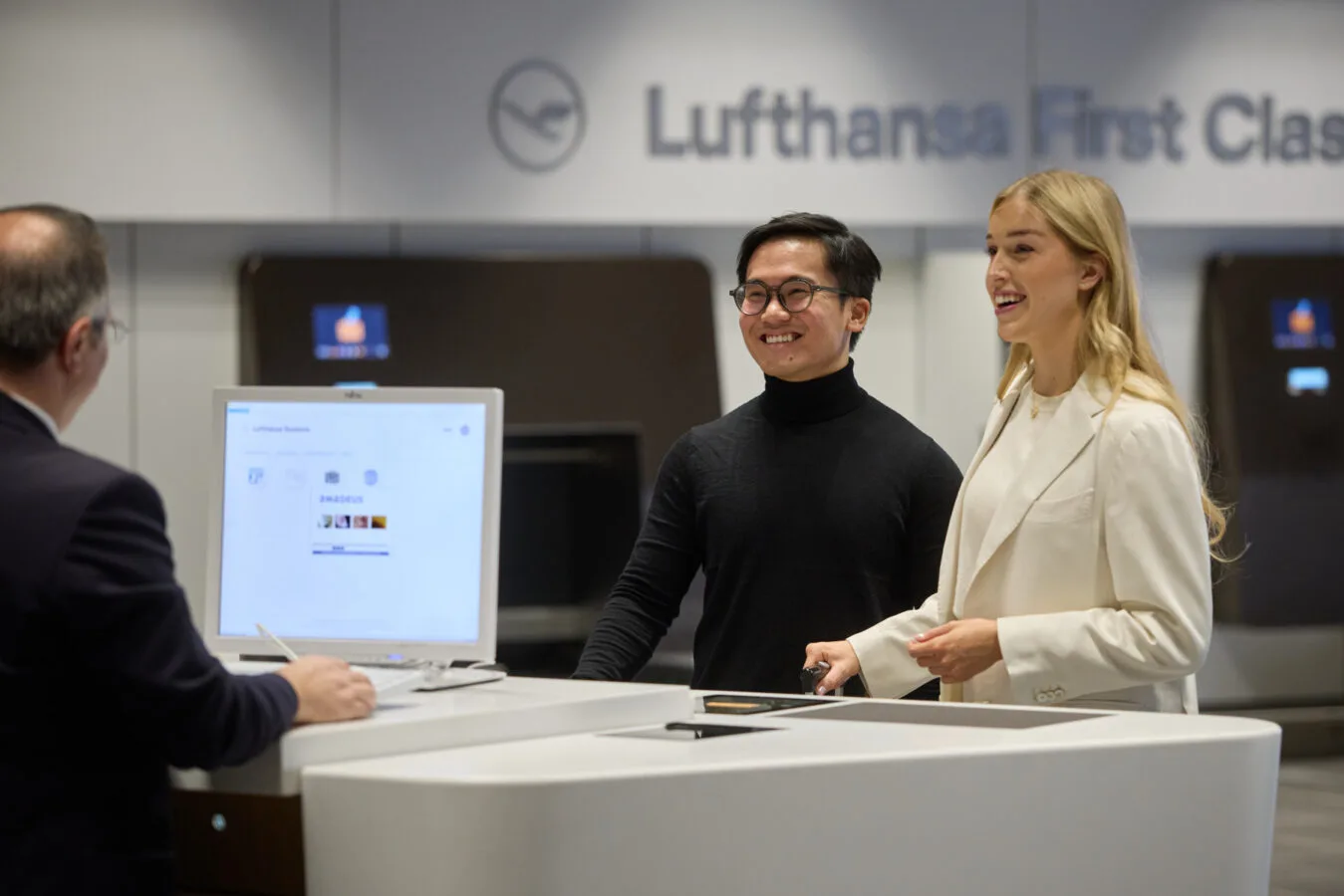 Zwei lächelnde Passagiere am Lufthansa First Class Schalter beim Premium Check-In Bereich, Mitarbeiter am Schalter und Check-in Monitor im Hintergrund