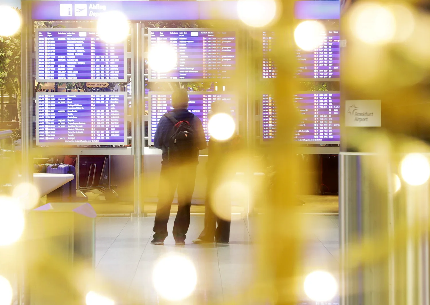 Passagiere vor Abflugtafel im Flughafen Frankfurt mit unscharfer Weihnachtsdekoration im Vordergrund, festliche Lichter und Spendendekoration sichtbar