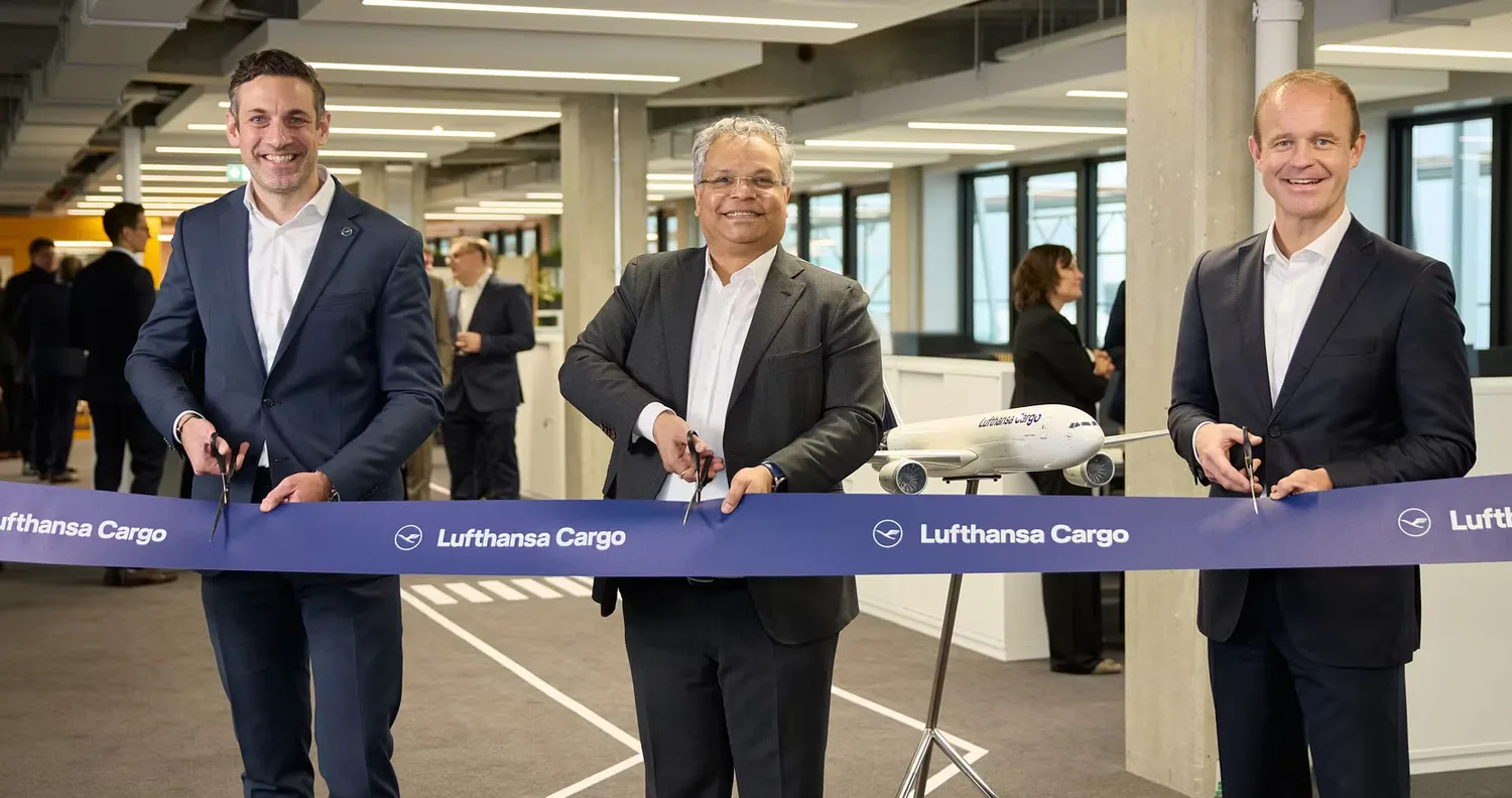 Drei Personen in Anzügen durchtrennen ein blaues Band mit Lufthansa Cargo Branding bei einer Eröffnungszeremonie in einem Büro von Fraport, Modellflugzeug im Hintergrund