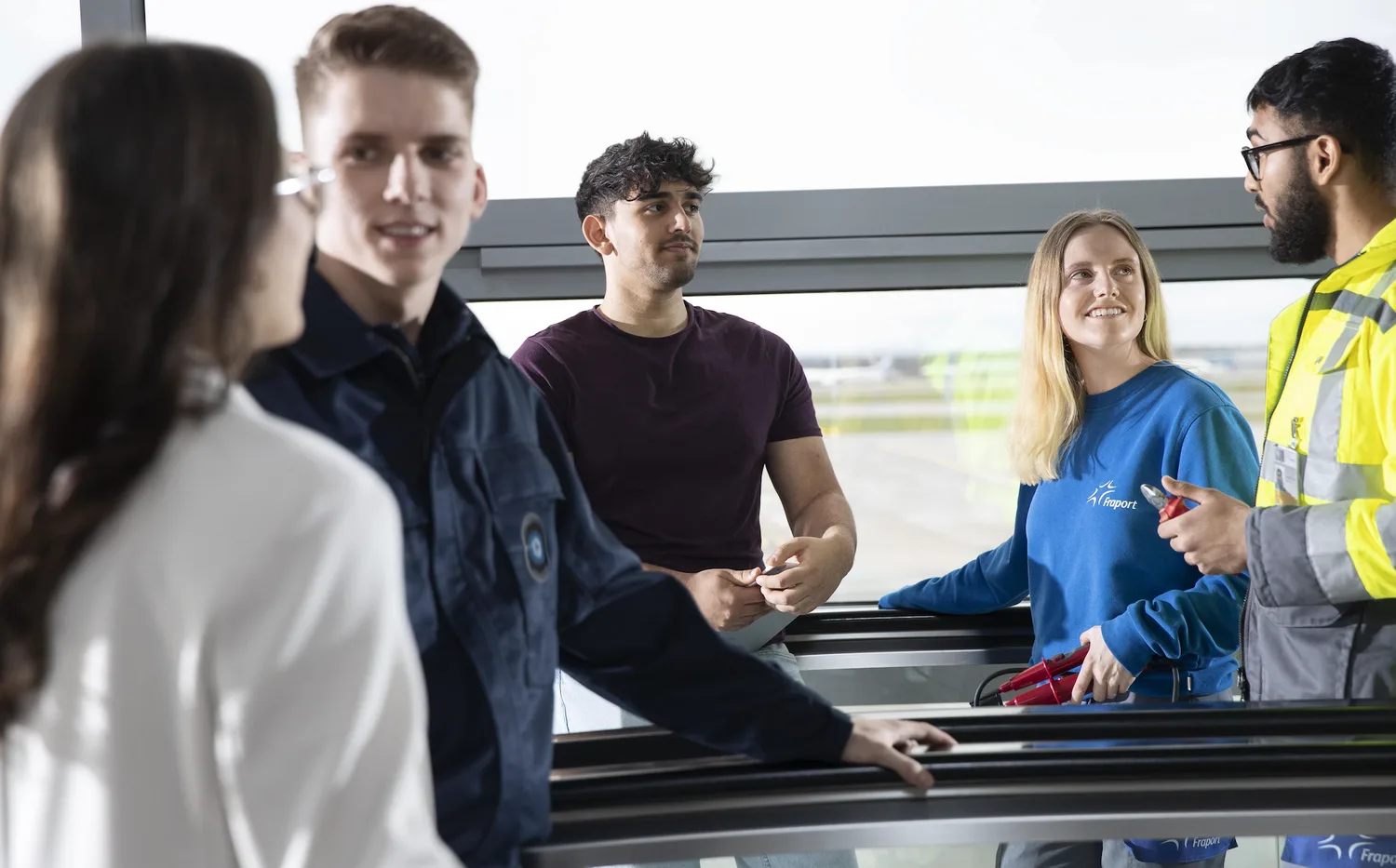 Gruppe junger Auszubildender am Flughafen in Gespräch, junge Frau mit Fraport-Pullover und Mitarbeiter in Sicherheitsweste
