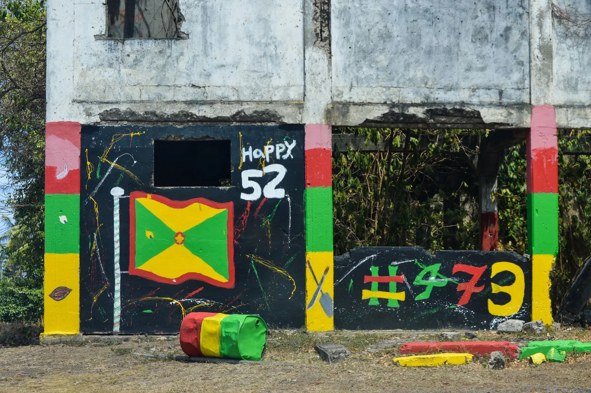 Verwitterte Hauswand mit bemalter Grenada-Flagge, Schriftzug Happy 52 und bunt gestrichenem Fass
