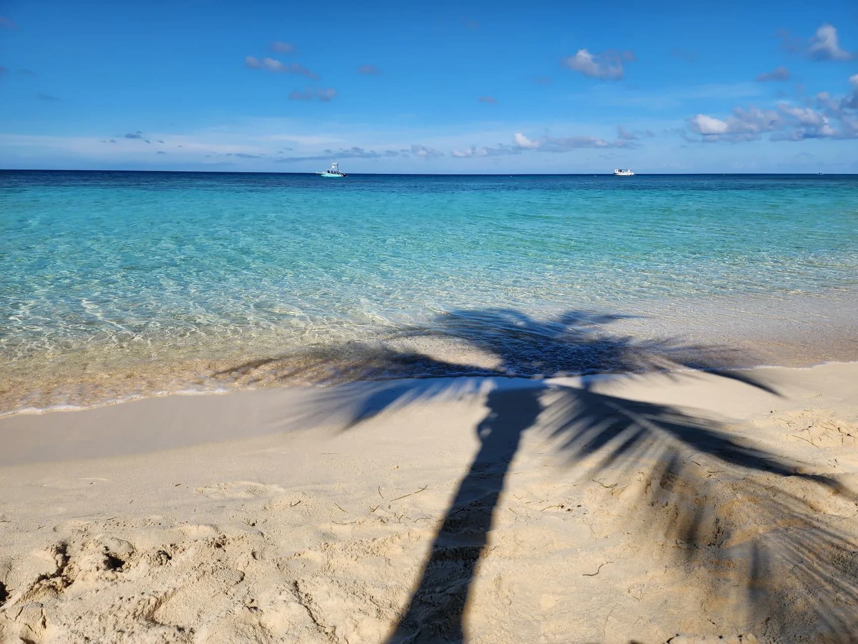 Schatten einer Palme auf weißem Sandstrand mit türkisfarbenem Meer und zwei Booten am Horizont
