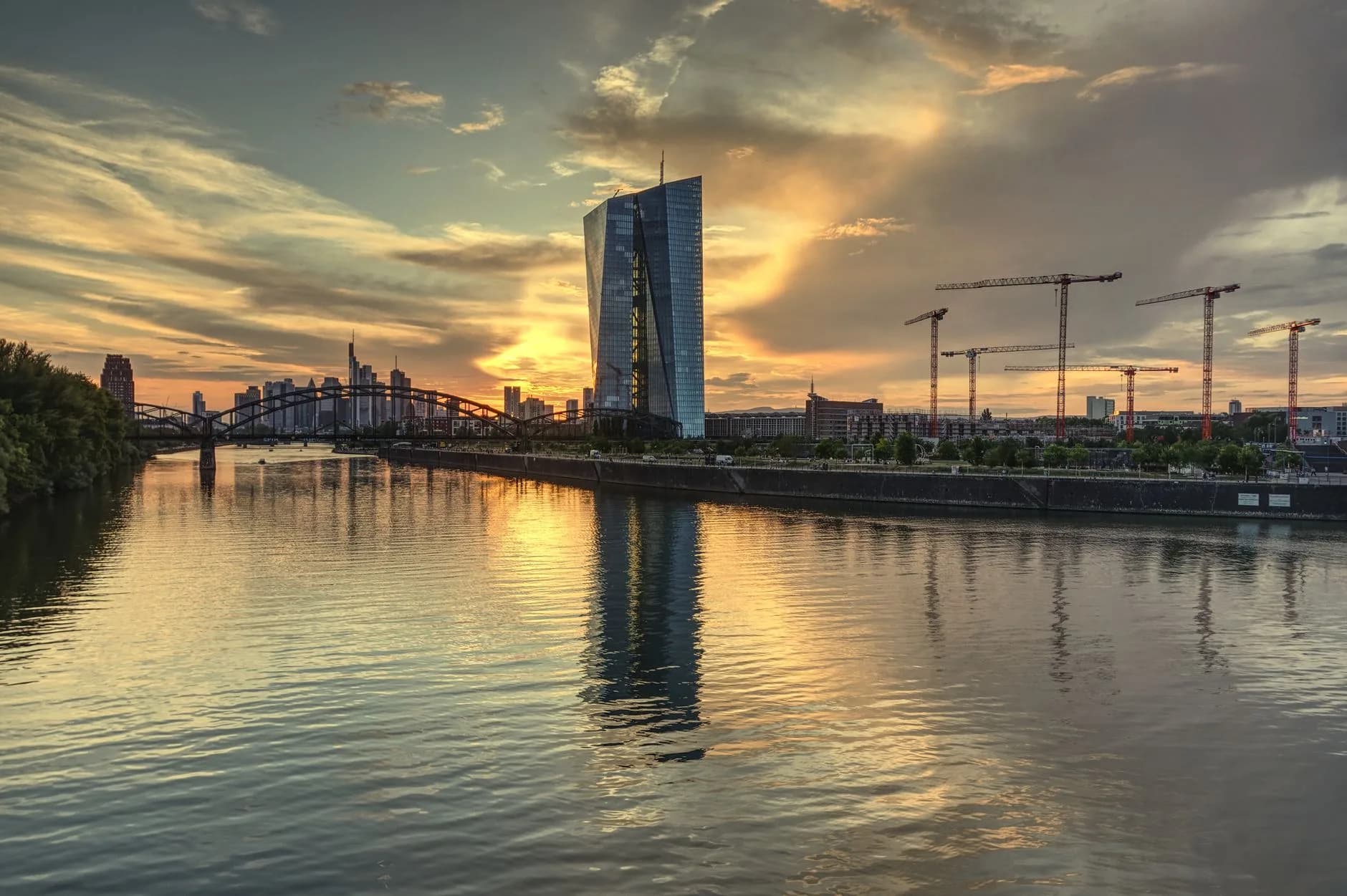 Hochhaus der EZB am Mainufer in Frankfurt bei Sonnenuntergang mit Baukränen und Skyline im Hintergrund