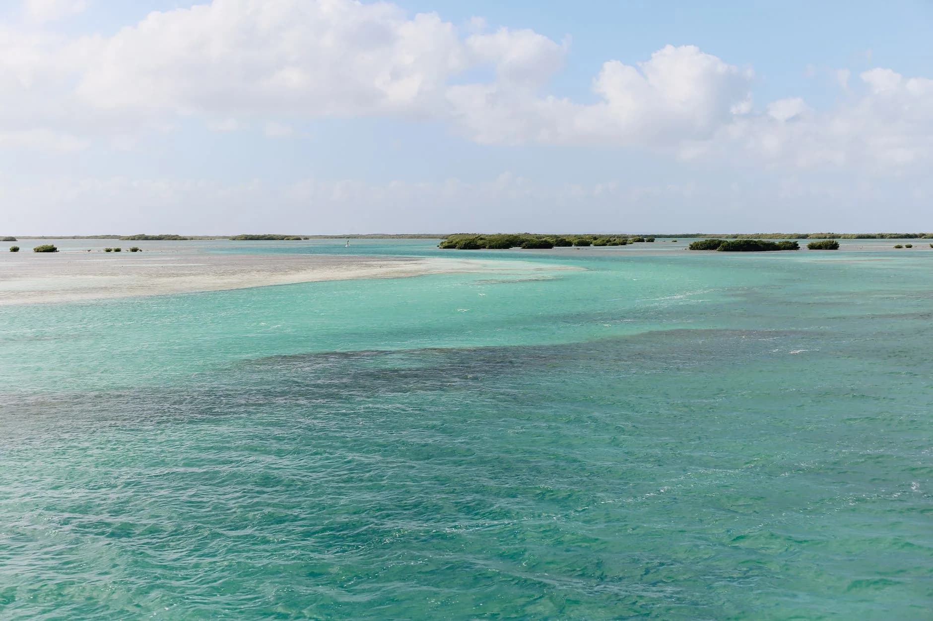 Türkisfarbenes Flachwasser mit Sandbänken und kleinen Mangroveninseln am Horizont unter Wolkenhimmel