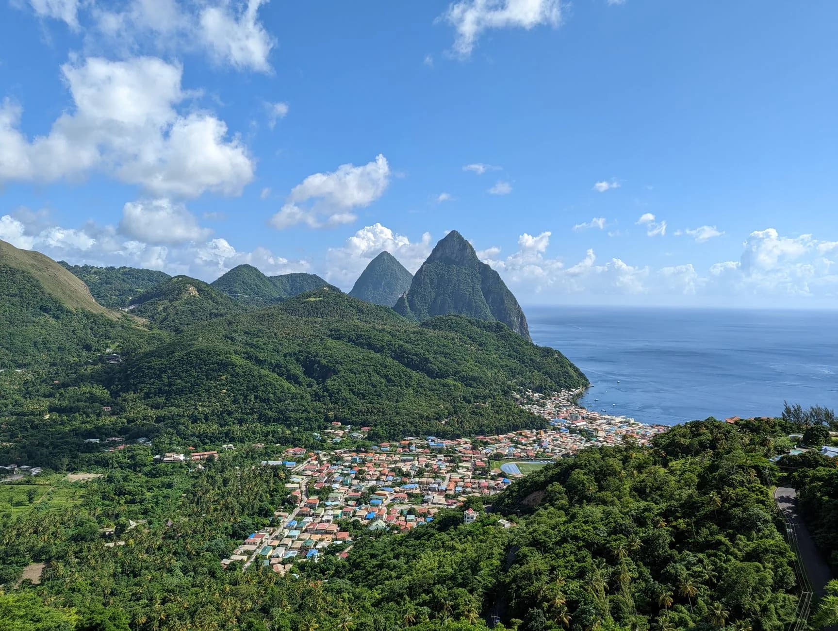 Blick über grüne Hügel auf eine Küstenstadt mit zwei markanten Felsgipfeln und dem Meer im Hintergrund