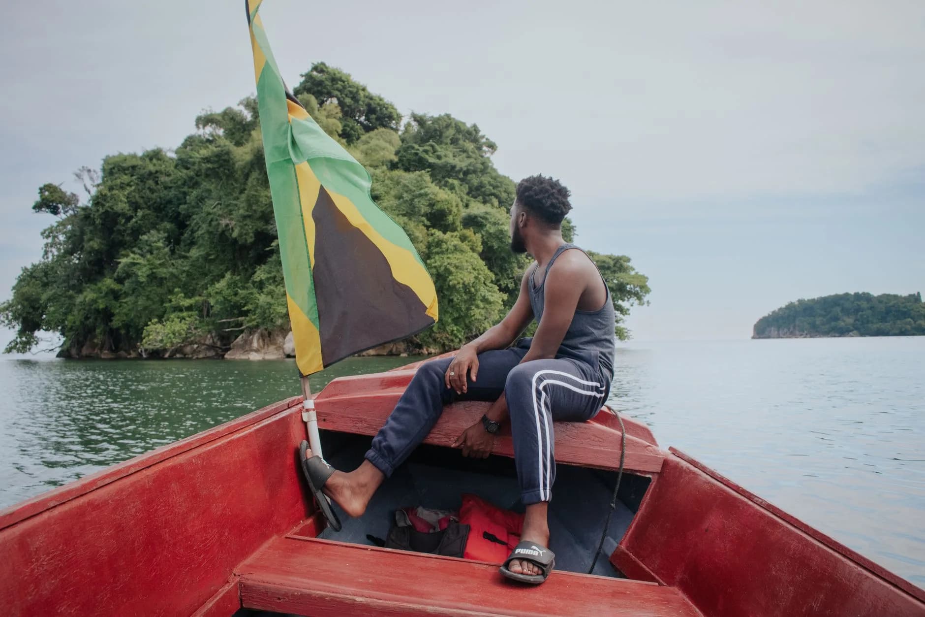 Mann sitzt am Bug eines roten Bootes mit jamaikanischer Flagge und blickt auf bewaldete Inseln im Wasser