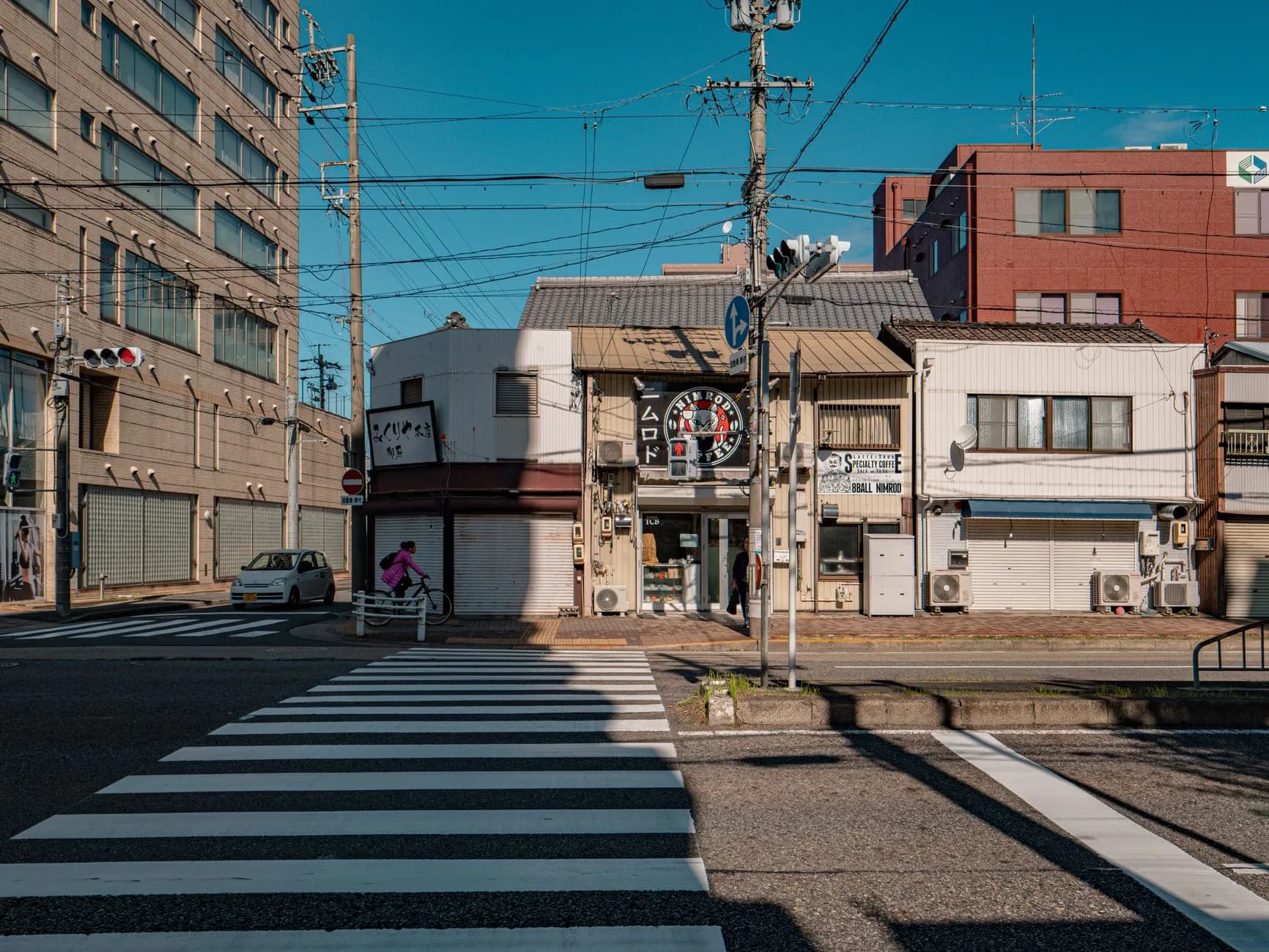 Japanische Straßenkreuzung mit Zebrastreifen, Radfahrerin und kleinen Ladengeschäften unter blauem Himmel