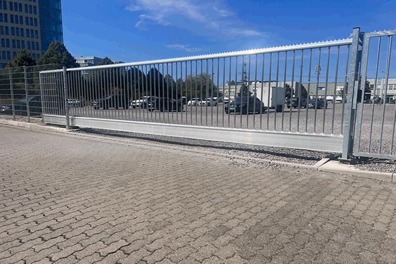 Metallschiebetor vor umzäuntem Parkplatz mit geparkten Autos und gepflasterter Zufahrt unter blauem Himmel.