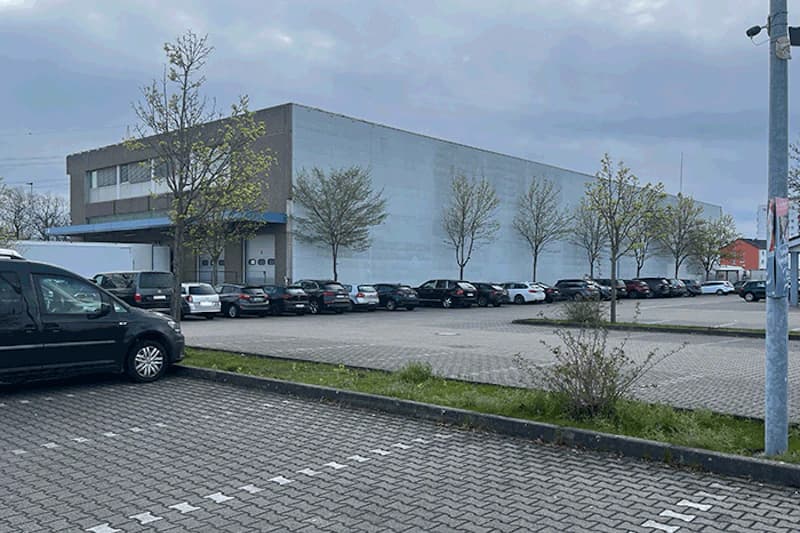 Großes Lagergebäude neben gepflastertem Parkplatz mit geparkten Autos, jungen Bäumen und Überwachungskamera auf Laterne.
