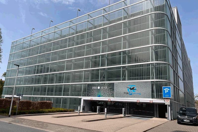 Mehrstöckiges Parkhaus mit durchlässiger Metallfassade, Einfahrt mit Schranken und blauem Parkzeichen, schwarzer Van rechts.