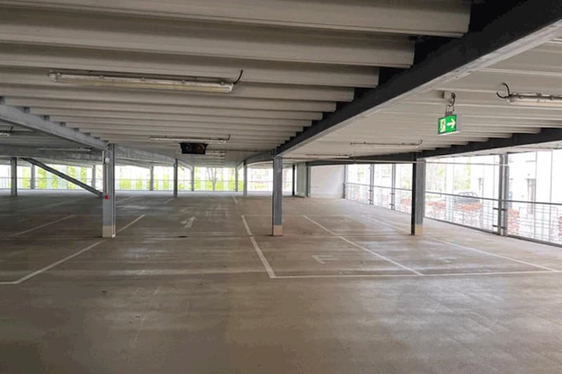 Leeres Parkdeck mit markierten Stellplätzen, Stahlstützen, Deckenbeleuchtung und Notausgangszeichen an der Seite.