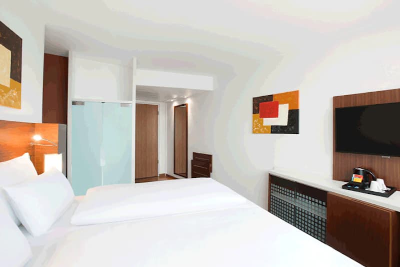 Hotelzimmer mit Doppelbett, Holzkopfteil, Fernseher an der Wand und Milchglas-Schiebetür zum Bad.