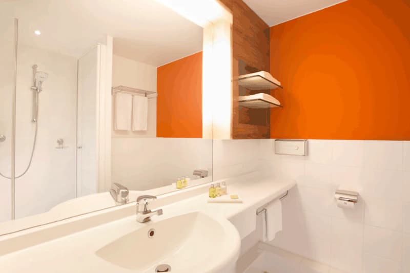 Badezimmer mit Waschbecken, großem Spiegel, oranger Wand, Duschkabine, Handtüchern und Pflegeartikeln.
