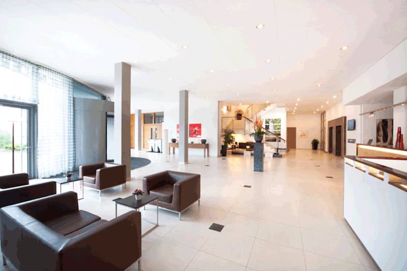 Helle Hotellobby mit braunen Ledersesseln, niedrigen Tischen, Rezeption rechts und Treppe im Hintergrund.