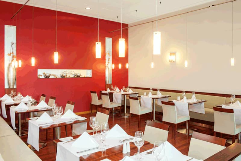 Hotelrestaurant mit gedeckten Tischen, weißen Servietten, hängenden Lampen und roter Akzentwand.