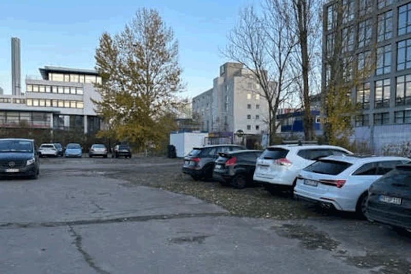 Offener Parkplatz mit mehreren geparkten Pkw auf rissigem Asphalt, kahlen Bäumen und mehrstöckigen Gebäuden im Hintergrund.
