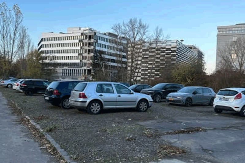 Freier Schotterparkplatz mit mehreren geparkten Autos vor mehrstöckigen Bürogebäuden und kahlen Bäumen.