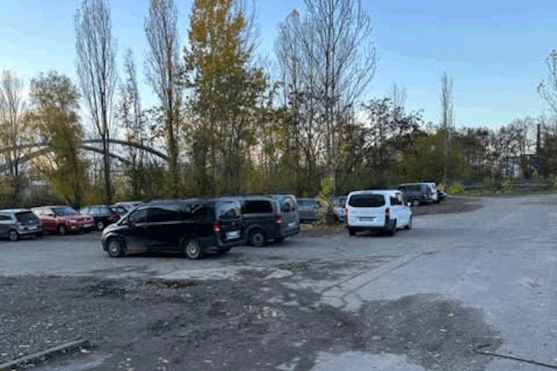 Parkplatz mit Schotter- und Asphaltfläche, mehreren geparkten Autos und Bäumen am Rand bei klarem Himmel.