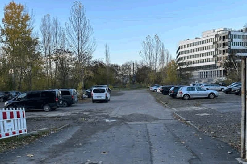 Großer Außenparkplatz mit geparkten Autos, rissigem Asphalt und kahlen Bäumen, Bürogebäude im Hintergrund.