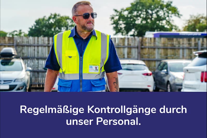 Mann mit gelber Warnweste und Sonnenbrille vor geparkten Autos und Holzzaun.