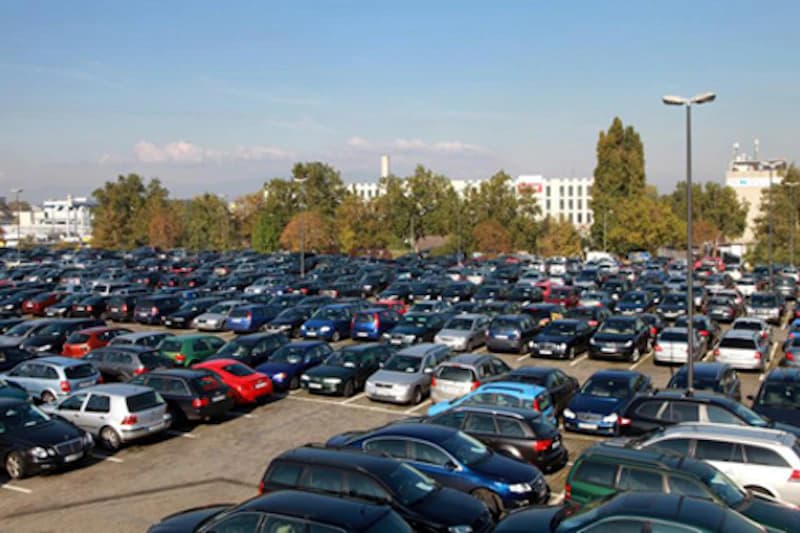 Außenparkplatz mit dicht geparkten Autos, Beleuchtungsmasten und Bäumen vor Gebäuden.