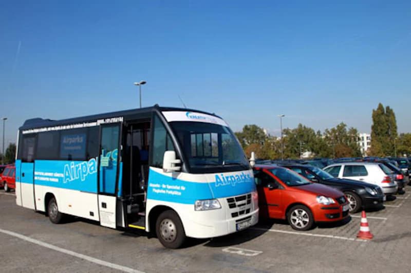 Weiß-blauer Airparks Transferbus mit offener Tür auf einem Außenparkplatz neben geparkten Autos und einem Verkehrshütchen bei klarem Himmel.