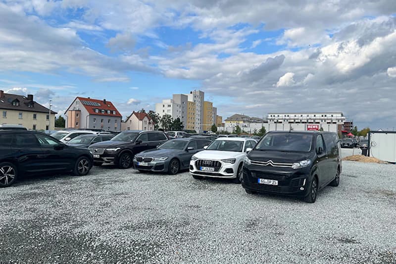 Parkplatz mit Schotter und mehreren geparkten Autos, Wohnhäusern und REWE im Hintergrund, bewölkter Himmel.