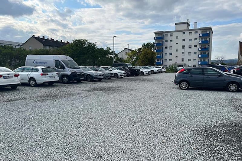 Schotterparkplatz mit Reihen geparkter Autos und einem weißen Lieferwagen, im Hintergrund ein Wohnhaus mit blauen Balkonen.