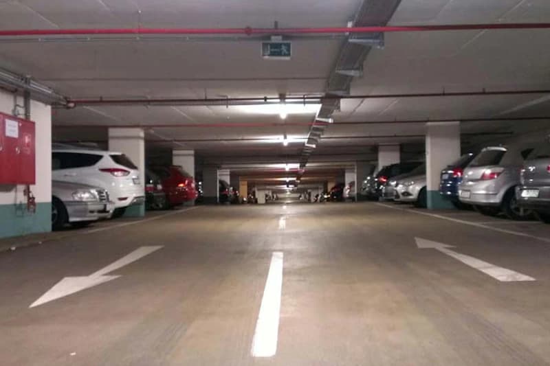 Tiefgarage mit geparkten Autos links und rechts, Deckenleuchten, Fluchtwegzeichen und Fahrpfeilen auf dem Boden.