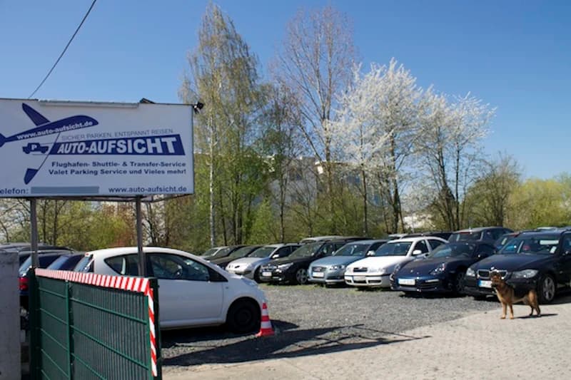 Geparkte Autos auf umzäuntem Parkplatz am Flughafen Frankfurt, Werbeschild Auto-Aufsicht und Hund.