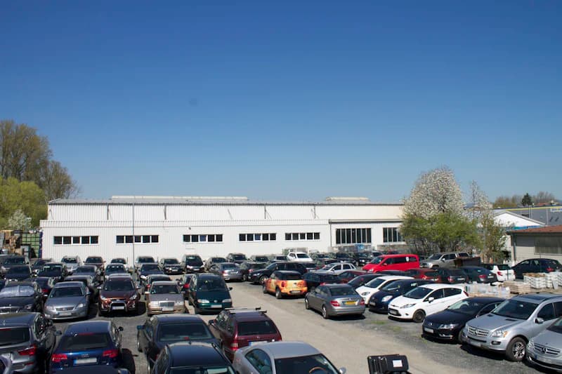 Voller Außenparkplatz mit zahlreichen geparkten Autos vor einer weißen Lagerhalle und blauem Himmel.