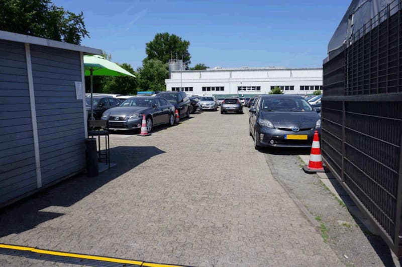 Gepflasterter Parkplatz mit geparkten Autos, Absperrkegeln, kleinem Häuschen und Gebäude im Hintergrund.