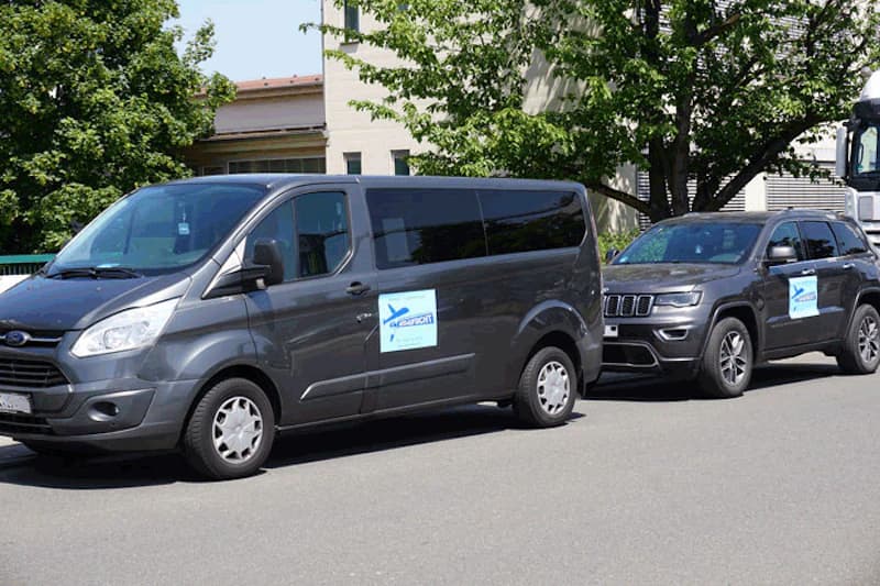 Zwei graue Fahrzeuge, ein Transporter und ein SUV, mit blauen Aufklebern stehen auf der Straße vor Gebäuden und Bäumen.