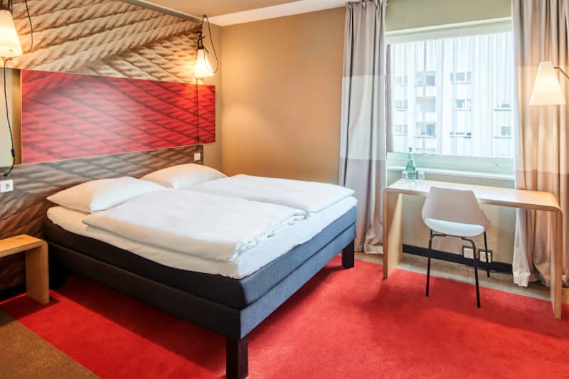 Zweibettzimmer mit zusammengestellten Betten, Nachttischlampen, Schreibtisch am Fenster und rotem Teppich.